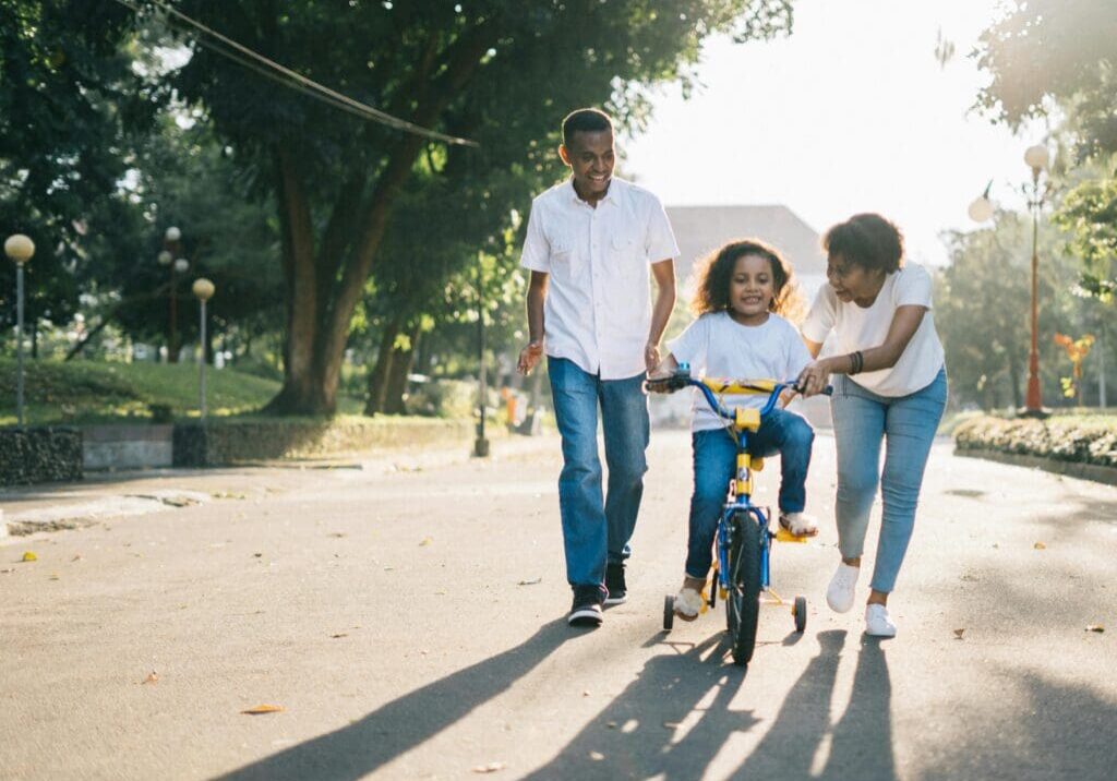 A young girl riding on the back of her bike while being pulled by an adult.